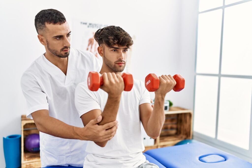 Two hispanic men physiotherapist and patient having rehab session using dumbbells at clinic